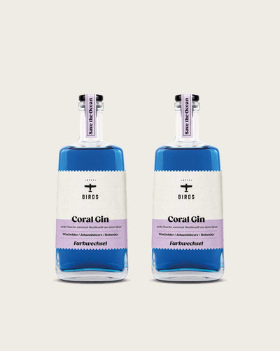 Zu sehen sind zwei Flaschen unseres Coral Gin, der Farbwechsel Gin mit Impact. Es handelt sich um zwei Glasflaschen, die blaue Farbe des Gins ist deutlich zu sehen. Das Etikett ist stilvoll in einem beige und lila gefärbt.