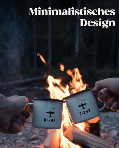 Mit zwei BIRDS Tassen wird vor einem Lagerfeuer angestoßen. Das Design der Tasse ist minimalistisch gehalten, auf der weißen Tasse befindet sich das schwarze Logo und der Markennamen BIRDS. 
